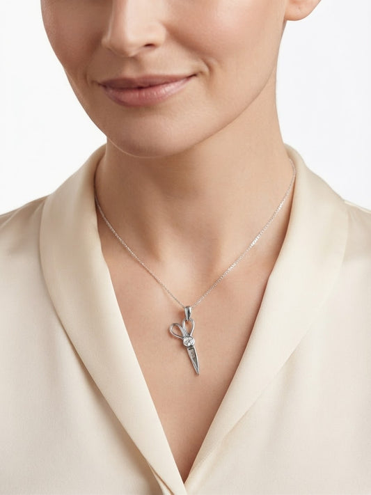 A close-up of a woman wearing a delicate silver necklace with a small pendant shaped like a pair of scissors, paired with a cream satin blouse against a light gray background.