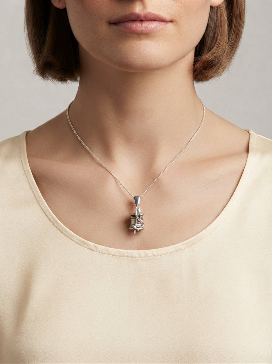 A close-up of a model wearing a delicate silver pendant necklace with a sculpted, textured pendant on a fine chain, paired with a cream-colored top against a neutral gray background.