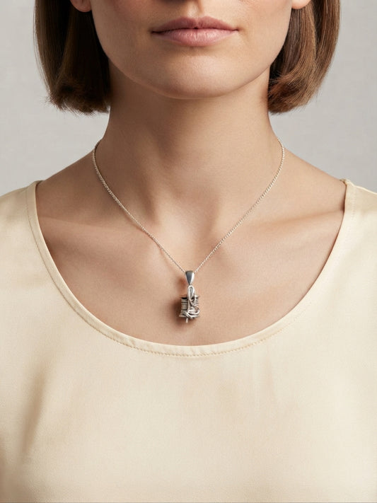A close-up of a model wearing a delicate silver pendant necklace with a sculpted, textured pendant on a fine chain, paired with a cream-colored top against a neutral gray background.