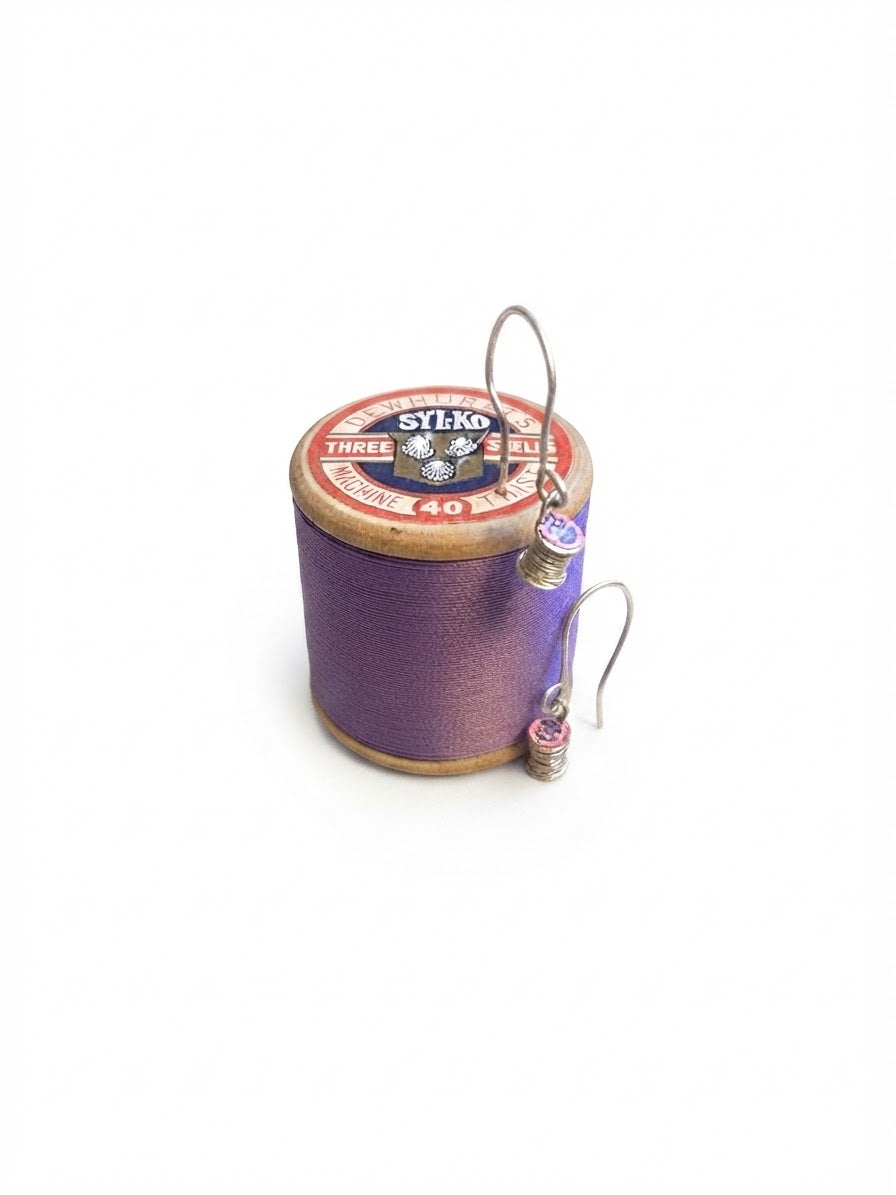 A pair of handmade silver hoop earrings with small purple and pink beads, draped over a vintage purple spool of thread on a white background.